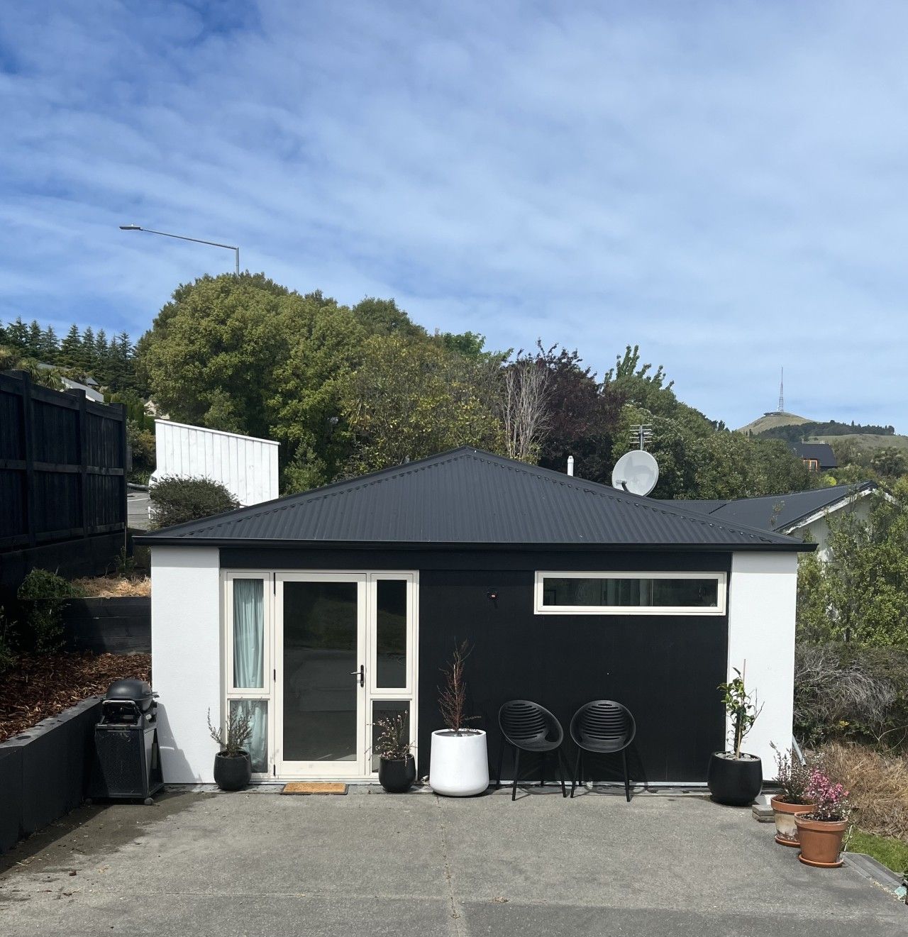 A modern single-story house with a black roof, white exterior walls, and a garage door. There are two potted plants and two chairs outside.