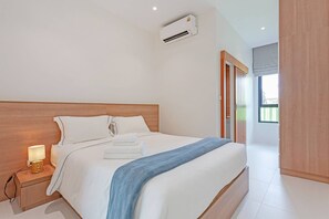 A modern bedroom with a wooden headboard, a bedside lamp, a neatly made bed with white linens, and an air conditioning unit above it. - Modern Pool Villa Near Bangtoa Beach 5 Mins (Choeng Thale)