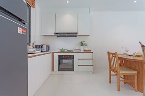A modern kitchen with white cabinetry, a built-in oven, and a dining area with a wooden table and chairs. - Modern Pool Villa Near Bangtoa Beach 5 Mins (Choeng Thale)