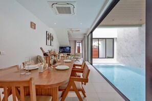 A modern dining area with a pool view, a television, and a ceiling fan. - Modern Pool Villa Near Bangtoa Beach 5 Mins (Choeng Thale)