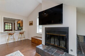 A living room with a fireplace, a television mounted above it, and a kitchen visible through an open doorway. - Plaza on Peacock Ave (Columbus)
