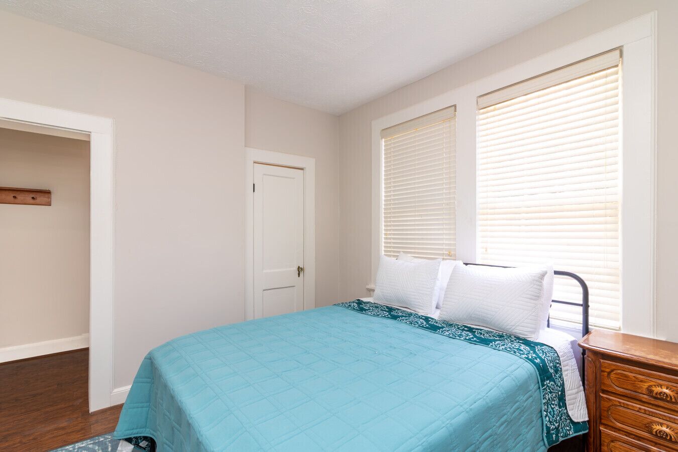 A bedroom with a bed, a dresser, a door, and a window with blinds.