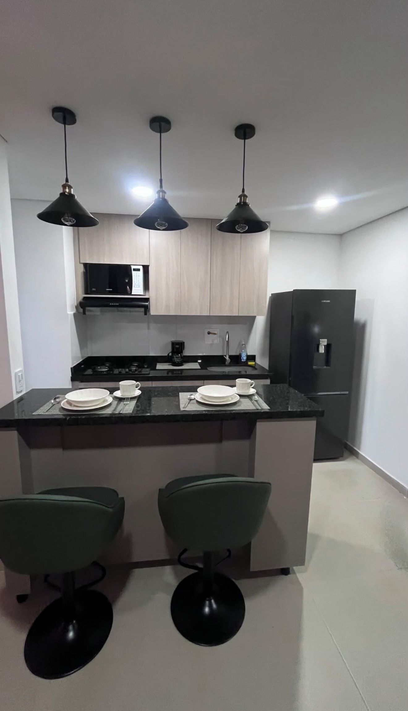 A modern kitchen with a central island, black pendant lights, and a refrigerator.