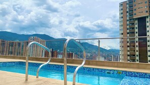 Piscina no terraço com grades de metal, borda em azulejo e paisagem urbana com montanhas ao fundo.