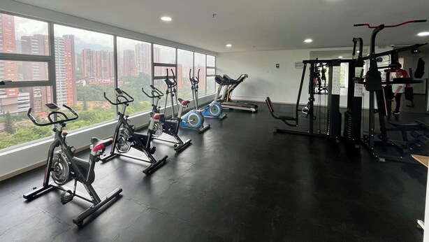 Uma academia com bicicletas ergométricas, esteiras e aparelhos de musculação, com amplas janelas e vista para a cidade.