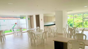 Uma ampla área de jantar com cadeiras e mesas de plástico branco, uma cozinha ao fundo e grandes janelas com vista para o verde.