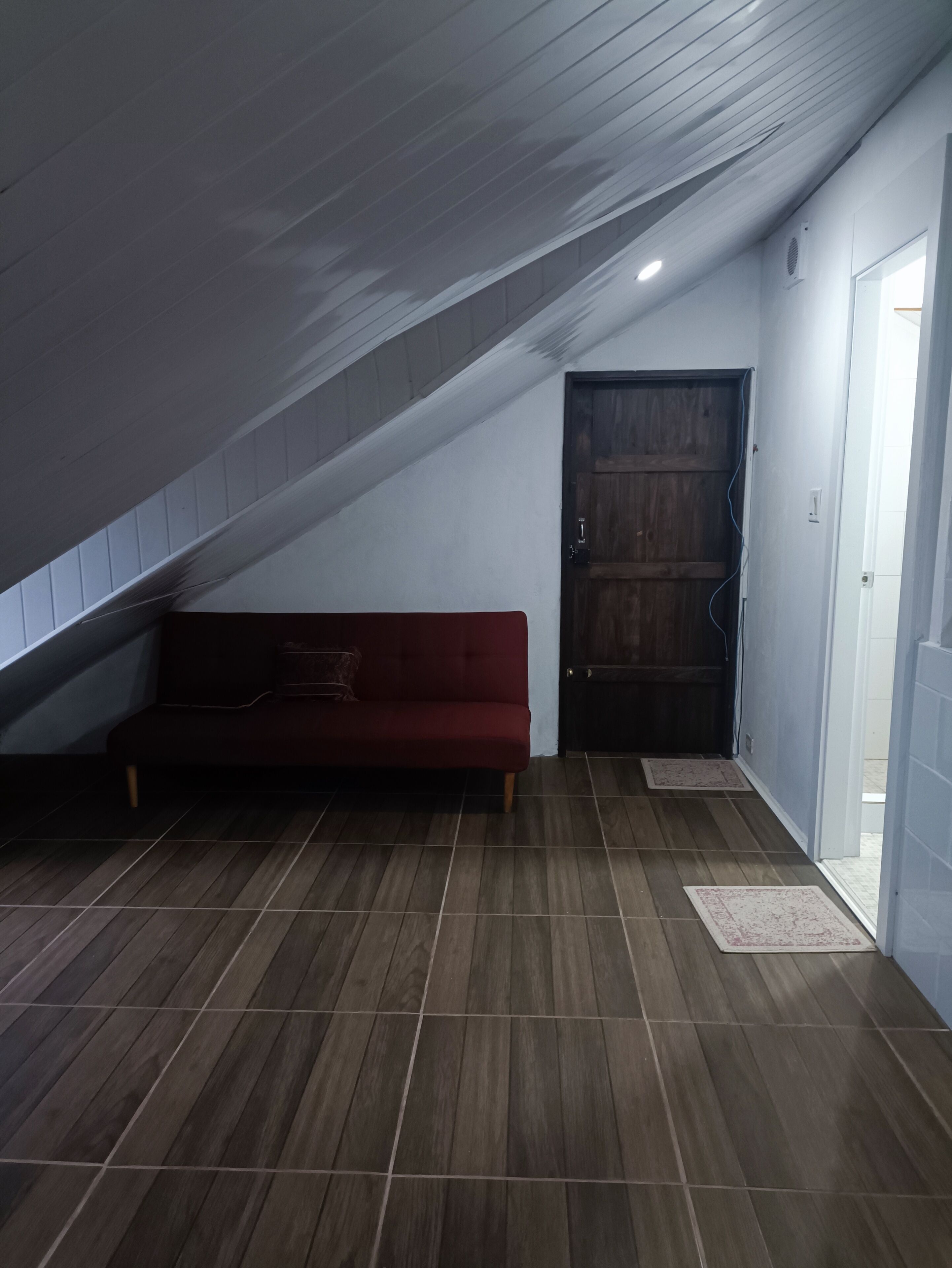 Una habitación con techo inclinado, una puerta de madera, un sofá rojo y suelo de baldosas.