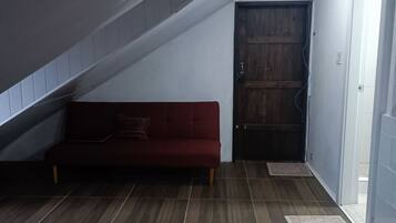 Una habitación con techo inclinado, una puerta de madera, un sofá rojo y suelo de baldosas.