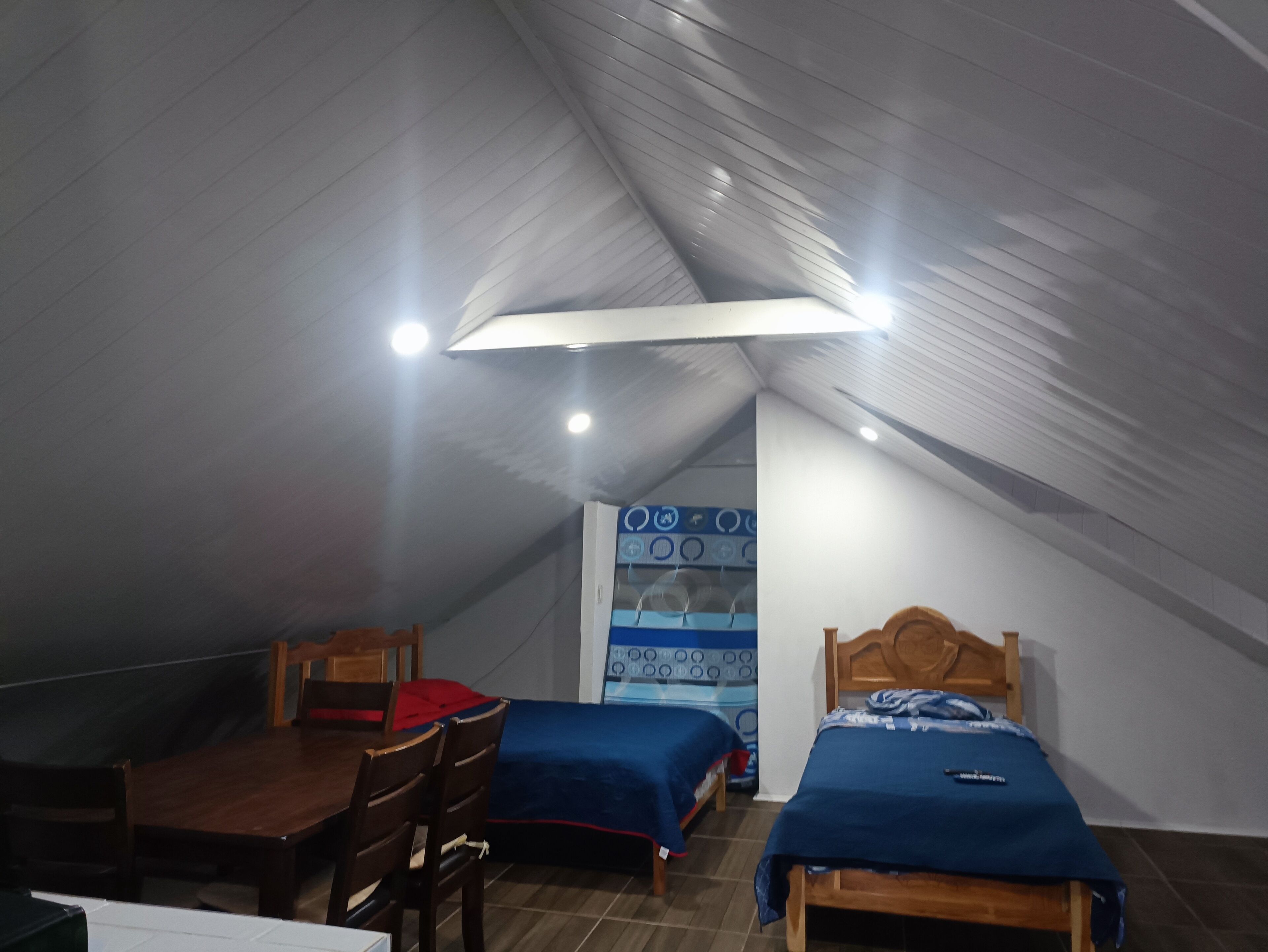 Ein kleiner Raum mit gedämpftem Licht, schrägem Dach, zwei Einzelbetten mit blauer Bettwäsche, einem Holztisch und einem Stuhl.