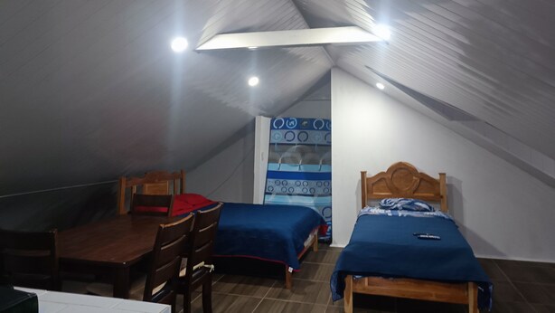 A small, dimly lit room with a sloped ceiling, two single beds with blue bedding, a wooden table, and a chair.