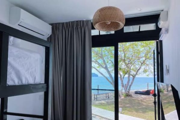 A room with a bed, a window with a view of the sea, and an air conditioning unit.