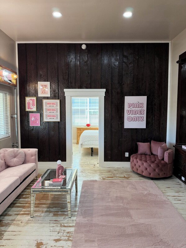 A room with a dark wooden wall, a pink sofa, a round pink chair, a glass coffee table, and a large pink rug.