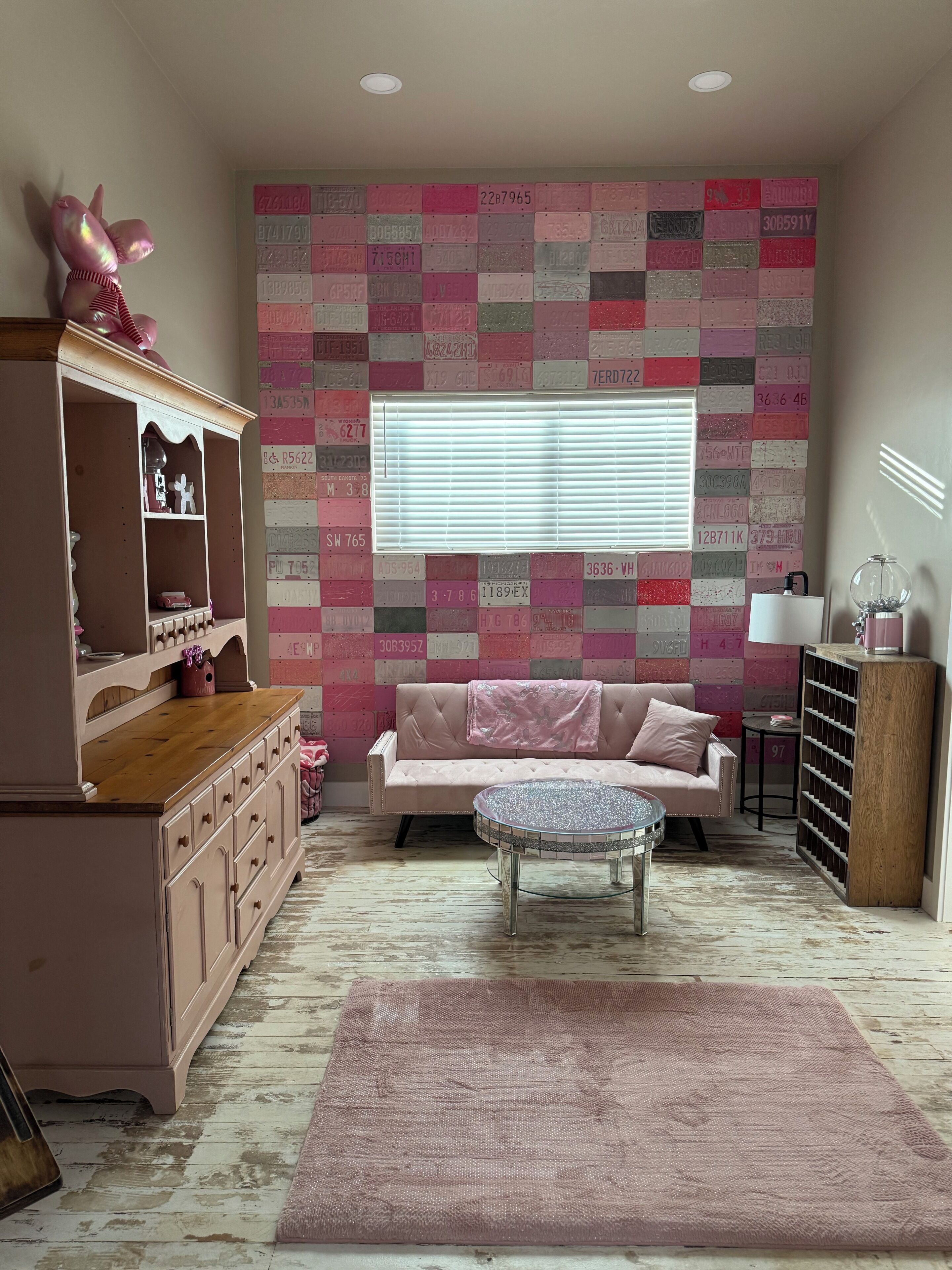 Een kamer met een roze bank, een rond glazen bijzettafeltje, een roze vloerkleed, een houten kast en een muur versierd met nummerborden.