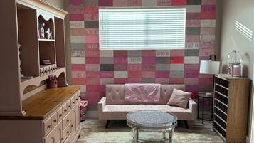 Een kamer met een roze bank, een rond glazen bijzettafeltje, een roze vloerkleed, een houten kast en een muur versierd met nummerborden.