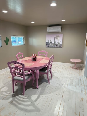 A room with a round table, pink chairs, a small pink table, a window with blinds, an air conditioning unit, and a wall-mounted picture.