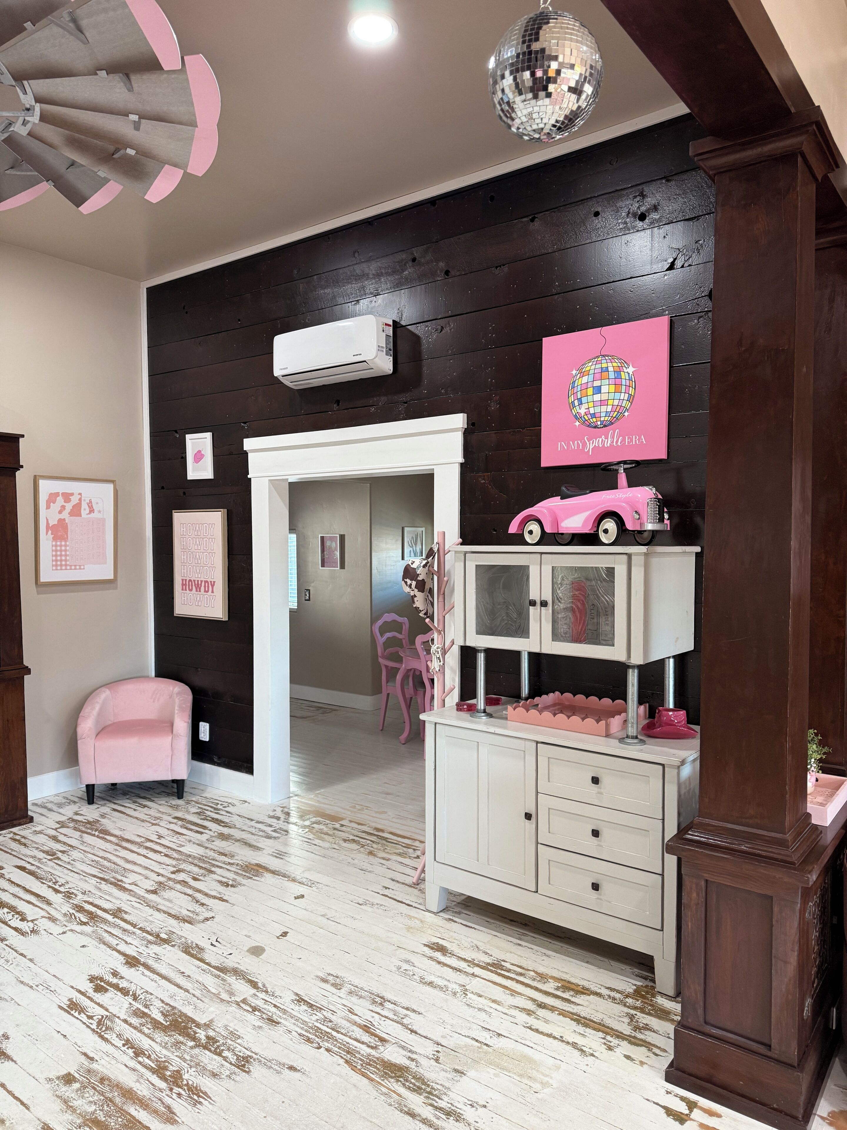 A room with a wooden ceiling, a disco ball, a pink toy car, and a white cabinet with drawers.