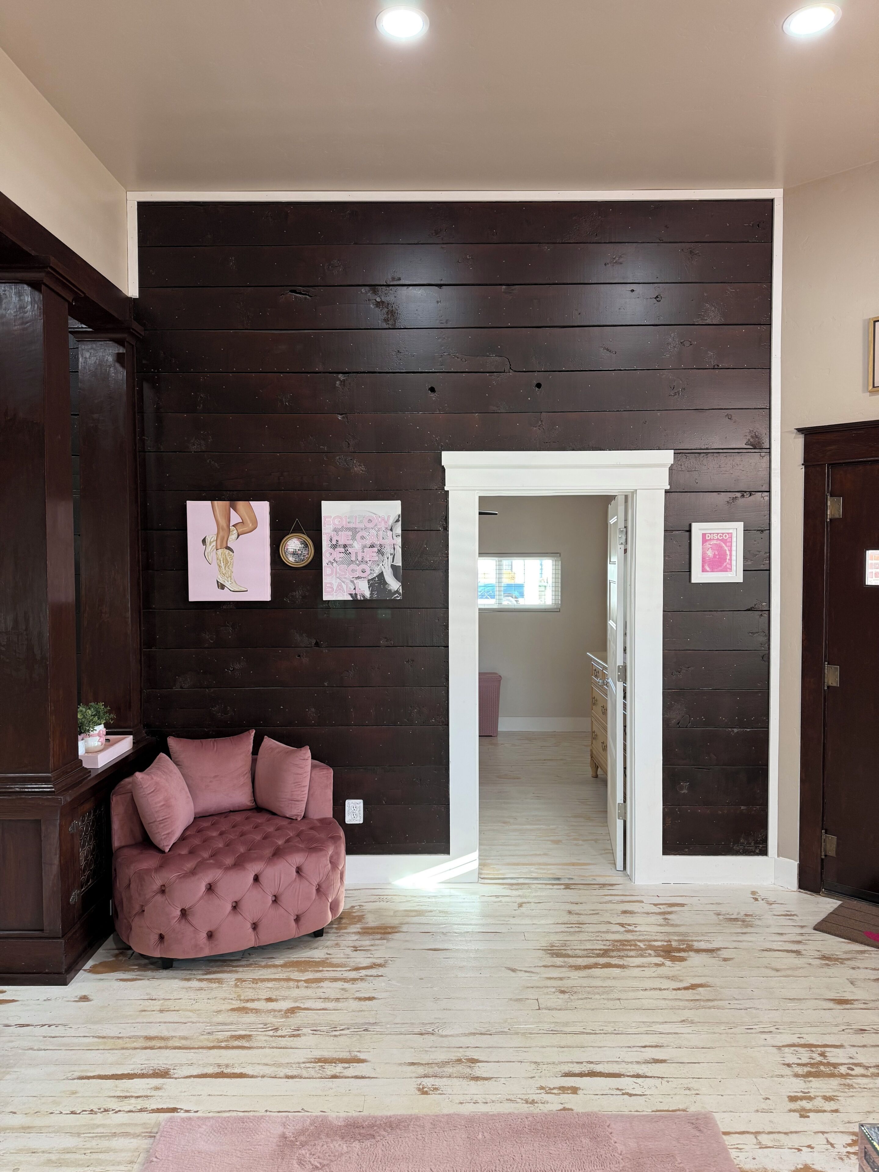 Una habitación con una pared de madera oscura, un sofá rosa con capitoné y una puerta blanca que da a otra habitación.