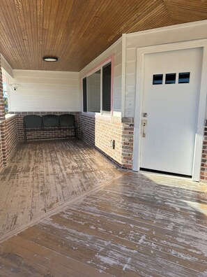 A covered patio area with a brick wall, a white door with three small windows, and a ceiling with wooden beams.