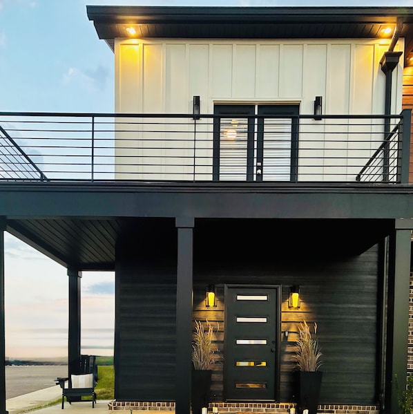 Una casa moderna de dos pisos con un balcón, una puerta principal con ventanal y un porche con una silla.