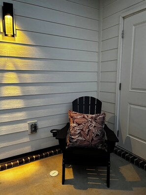 Eine Ecke eines Raumes mit einem schwarzen Adirondack-Sessel, einem gemusterten Kissen, einer Wandleuchte und einer Tür.
