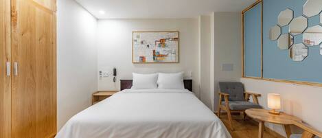 A hotel room with a large bed, a wooden wardrobe, a desk with a chair, a bedside table with a lamp, and a wall art piece.