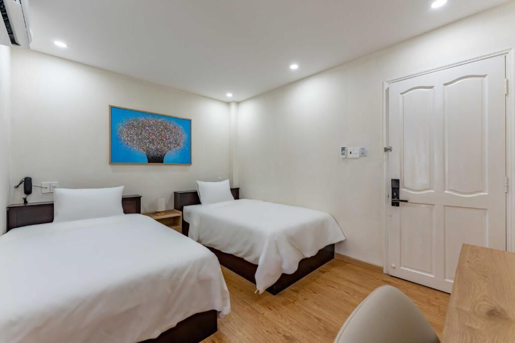A hotel room with two beds, a wooden floor, a white door, and a painting on the wall.