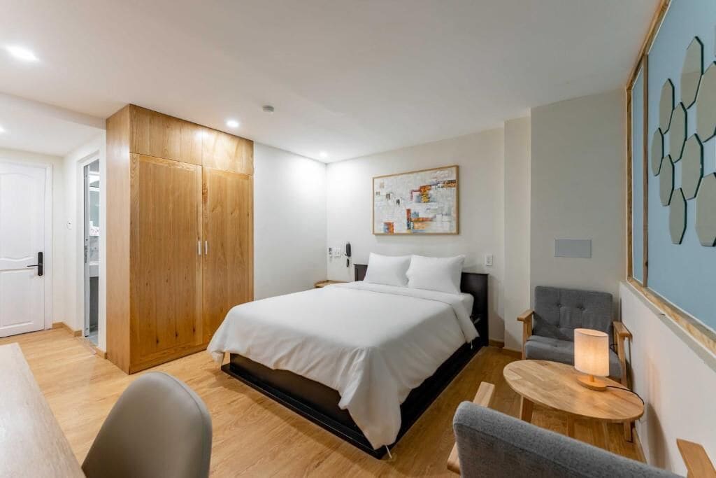 A hotel room with a bed, a desk, a chair, a wardrobe, and a wall with a geometric art piece.