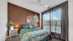 A bedroom with a bed, a nightstand, a potted plant, a wall art piece, and a window with curtains. - Coronation Station (Alpine)