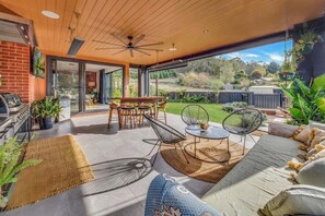A modern outdoor patio with a wooden ceiling, a dining table with chairs, and a seating area with cushions and a round table. - Coronation Station (Alpine)