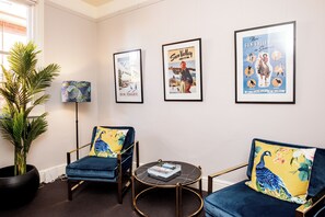 Ein Zimmer mit zwei blauen Sesseln, einem kleinen runden Tisch und einer Topfpflanze. An der Wand hängen drei gerahmte Poster.