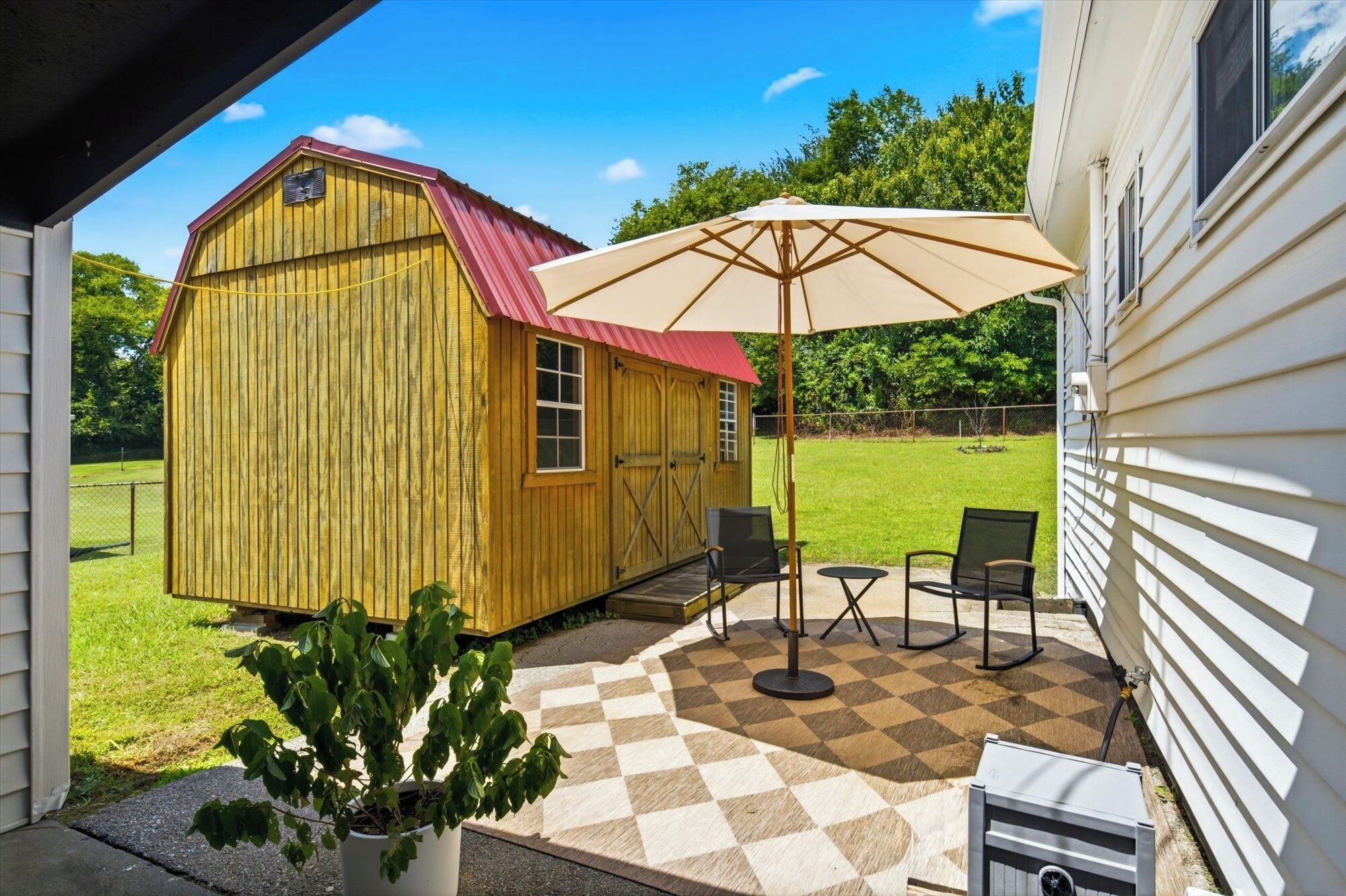 Een houten schuur met een rood dak, een terras met een ruitpatroon en twee stoelen, en een grote parasol.