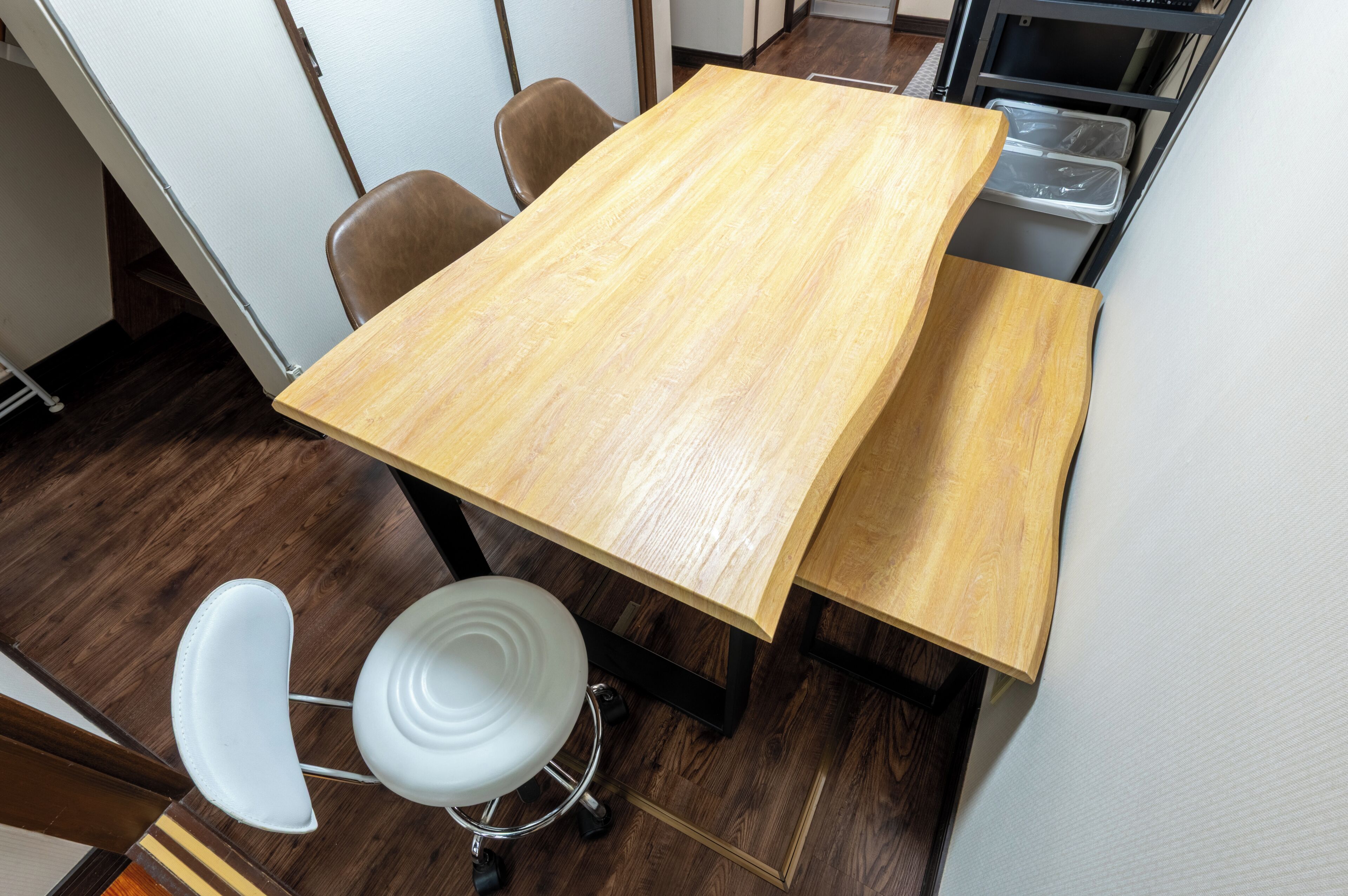 Een houten tafel met vier stoelen, een witte kruk en een zwart metalen rek met plastic bakken.