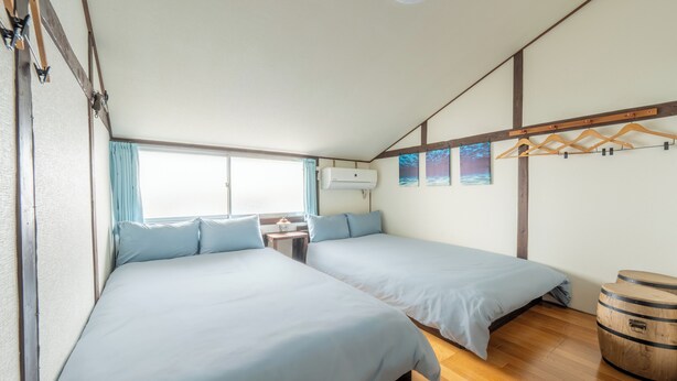 Un dormitorio con dos camas, suelo de madera, ventana con cortinas, aire acondicionado de pared y perchero de madera.