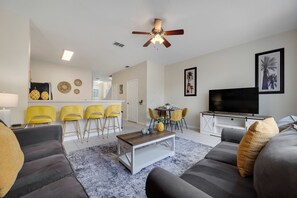 A modern living room with a sofa, coffee table, and a TV. There is a dining area with yellow chairs and a table. The room has a ceiling fan and artwork on the walls. - WP2355 - 3BR Townhome | Resort Access | Near Parks (Kissimmee)