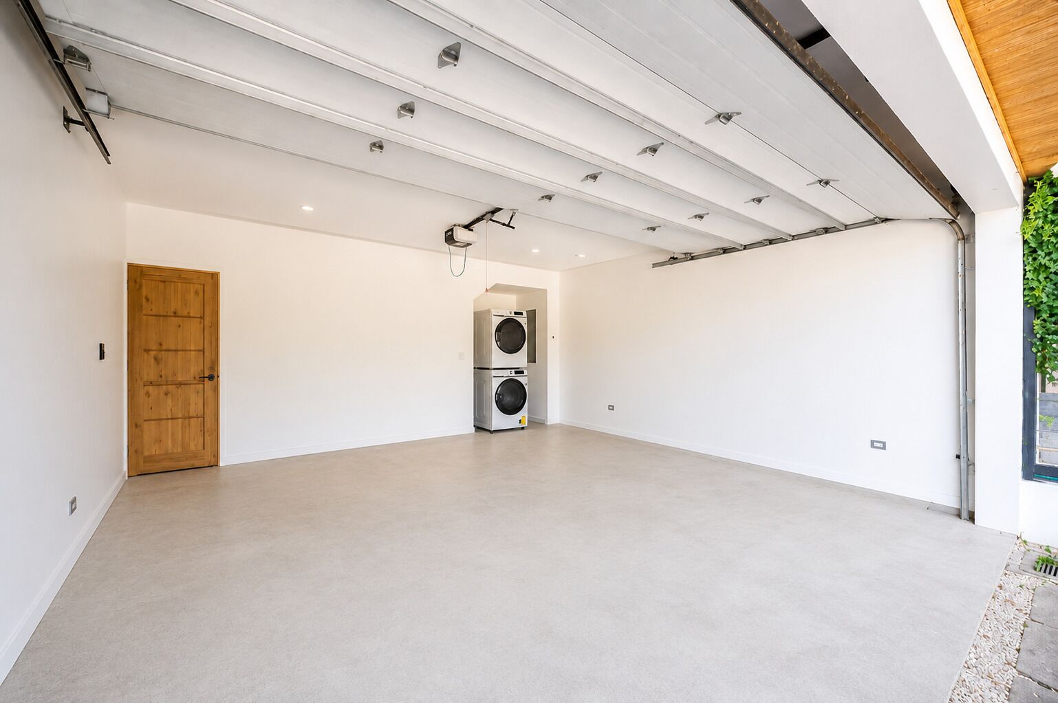 A spacious room with a large door, a washing machine, and a ceiling with exposed beams and lighting fixtures.