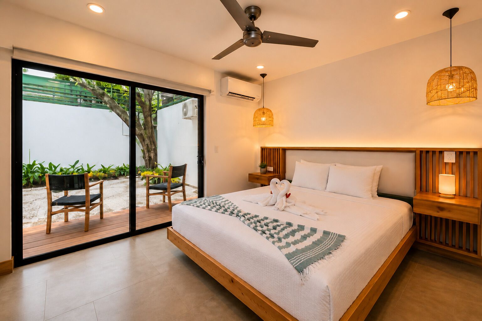 Habitación de hotel moderna con una cama grande, ventilador de techo, unidad de aire acondicionado, mesita de noche de madera, lámpara de noche y vistas a un espacio exterior con una silla y plantas.