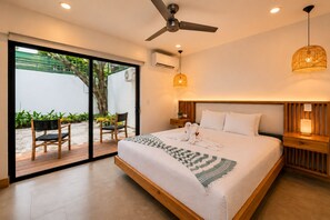 Ein modernes Hotelzimmer mit einem großen Bett, einem Deckenventilator, einer Klimaanlage, einem Nachttisch aus Holz, einer Nachttischlampe und Blick auf einen Außenbereich mit Stuhl und Pflanzen.