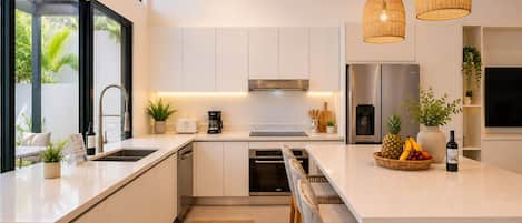 A modern kitchen with white cabinetry, a stainless steel refrigerator, a built-in oven, and a large island with a fruit bowl and wine bottle.