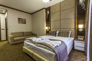 A hotel room with a large bed, a sofa, a bedside table with a lamp, and a wall-mounted artwork.