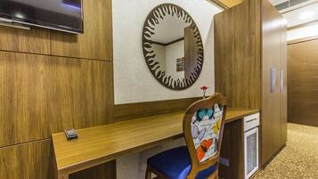 A hotel room with a wooden desk, a chair with a butterfly pattern, a small refrigerator, and a television mounted on the wall.
