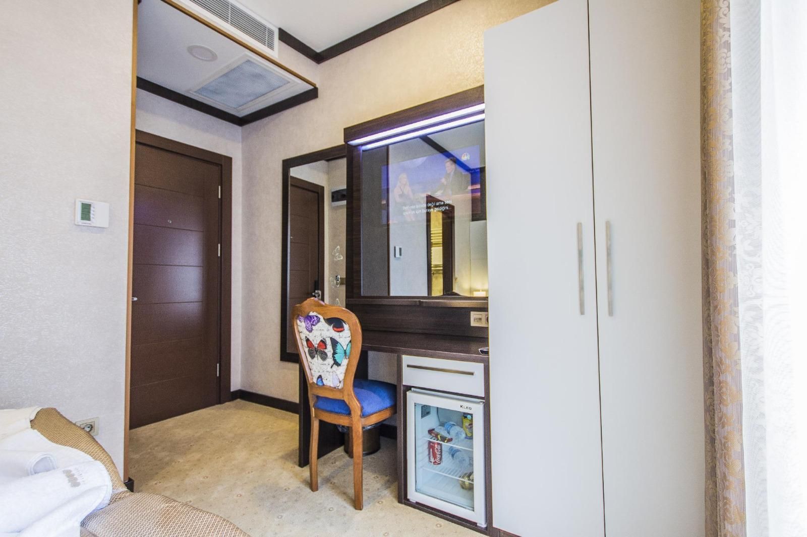 A hotel room with a wooden door, a chair with a colorful cushion, a small refrigerator, and a mirror above a vanity.
