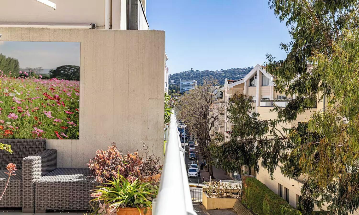 A balcony with wicker furniture, a flower bed, and a view of a city street.