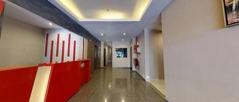 A hotel corridor with a red reception desk, a fire extinguisher, and a ceiling with recessed lighting.