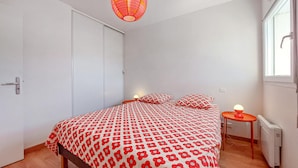 A bedroom with a bed, pillows, a nightstand, a window, and a radiator. - Charmant appart. proche plage (Royan)