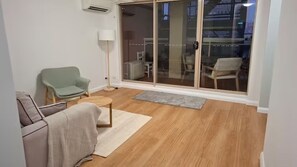 Ein Wohnzimmer mit einer Couch, einem kleinen Tisch, einer Stehlampe und einer Glastür, die auf einen Balkon mit Gartenmöbeln führt.
