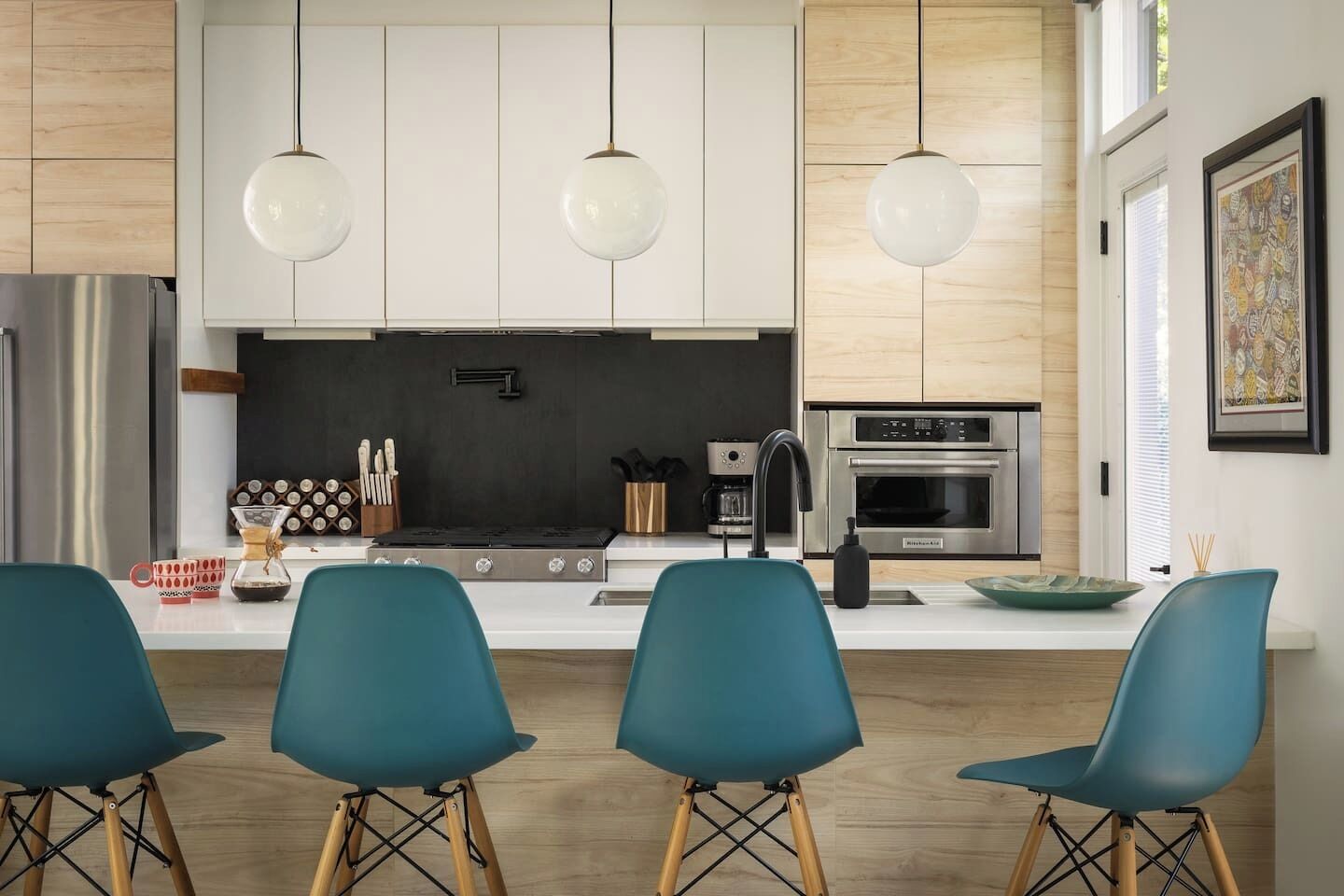 Una cocina moderna con armarios blancos, salpicadero negro, electrodomésticos de acero inoxidable, sillas color turquesa e iluminación colgante.