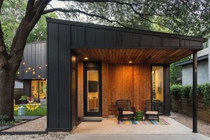 A modern outdoor seating area with two chairs and a small table, a wooden pergola, and a black exterior wall. - Modern Villa | Heated Pool + Movie + 6th St/DT (Austin)
