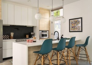 A modern kitchen with white cabinetry, a central island, and teal bar stools. - Modern Villa | Heated Pool + Movie + 6th St/DT (Austin)