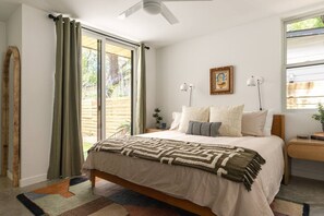 Ein Schlafzimmer mit Bett, Nachttisch, einem Fenster mit Vorhängen, Deckenventilator und einem gerahmten Bild an der Wand.
