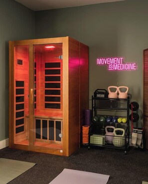 Ein Raum mit einer Holzsauna, einem leuchtenden Schild mit der Aufschrift 'MOVEMENT IS MEDICINE' sowie einem Gestell mit Kurzhanteln und Gewichten.